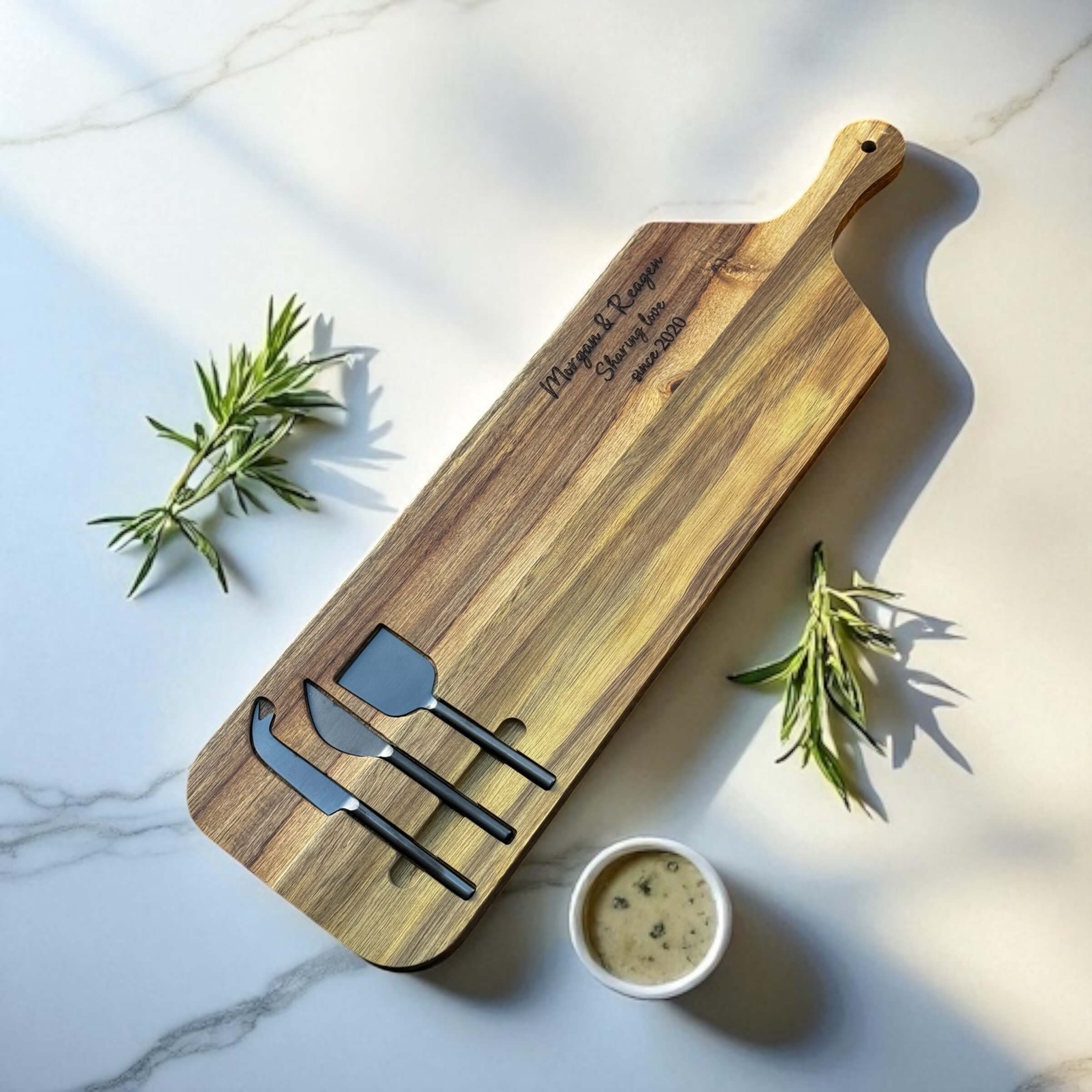 Elegant wooden cheeseboard featuring names and romantic engraving, styled with herbs and a dipping sauce.