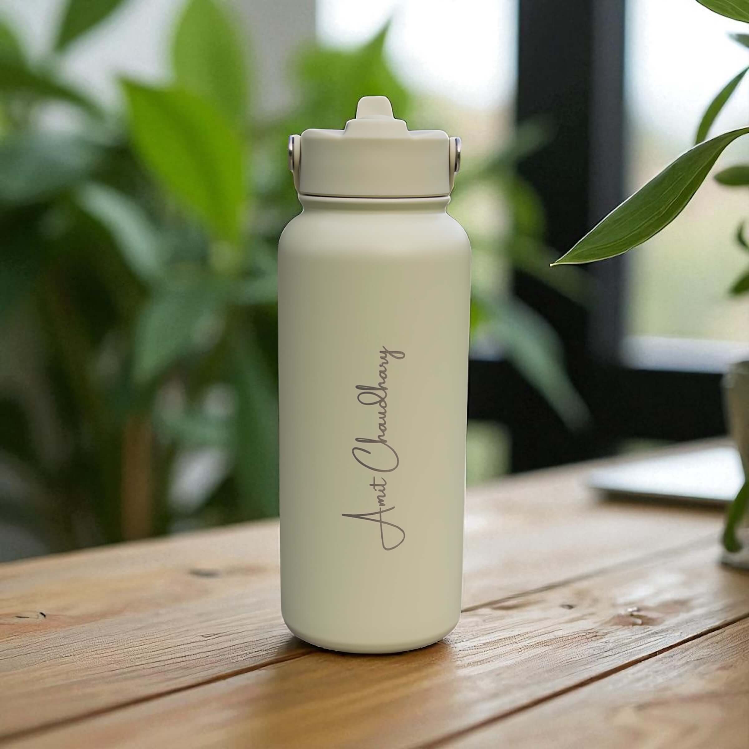 Sage 1L stainless steel water bottle personalised with engraved name, displayed on a wooden table with plants in the background.