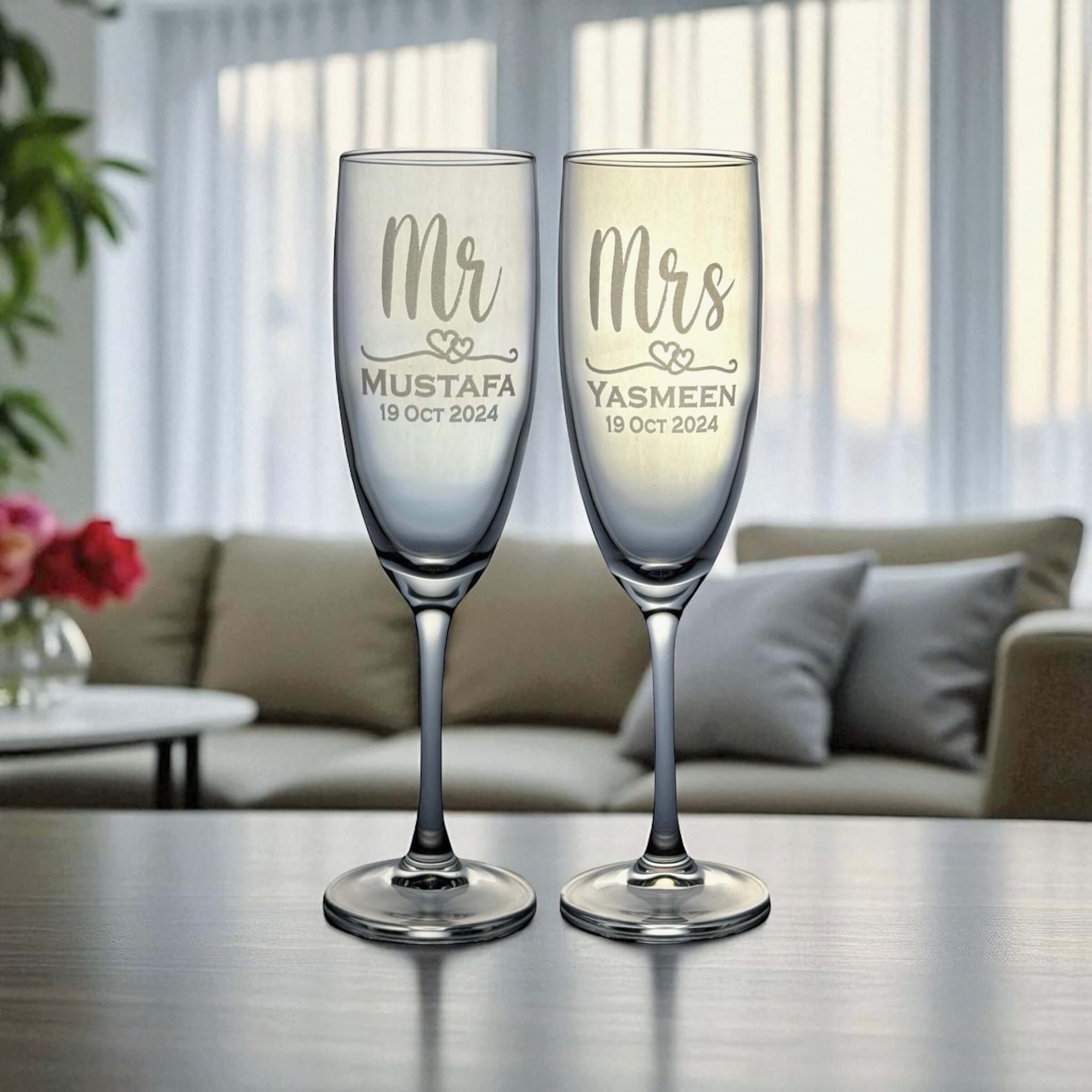 Two champagne flutes with 'Mr' and 'Mrs' , name and date engravings on a table in a living room setting.