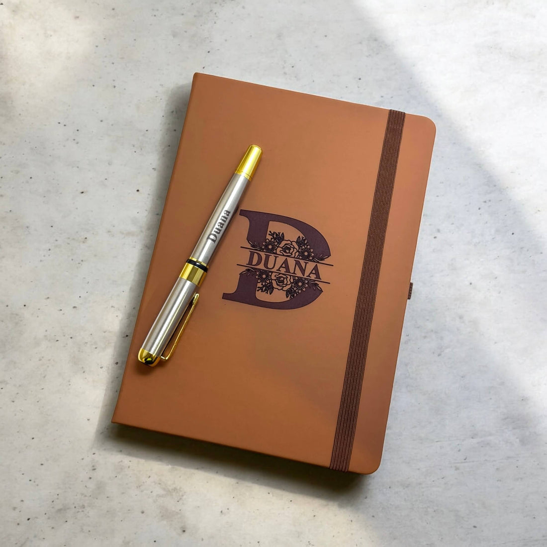 Personalised notebook with a split floral initial and engraved pen placed across the cover