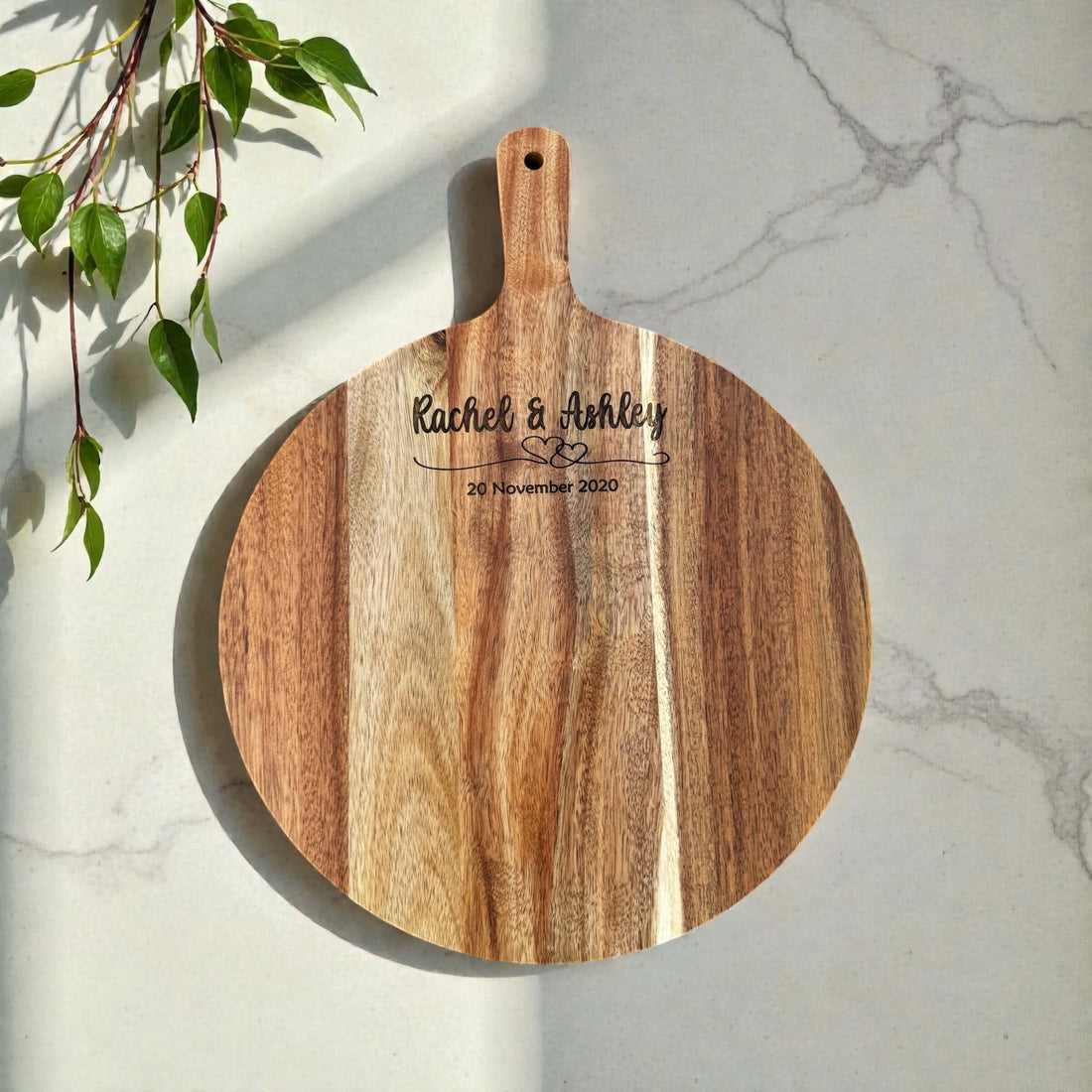 Round acacia serving board engraved with Rachel & Ashley and 20 November 2020, photographed on a marble surface with greenery.