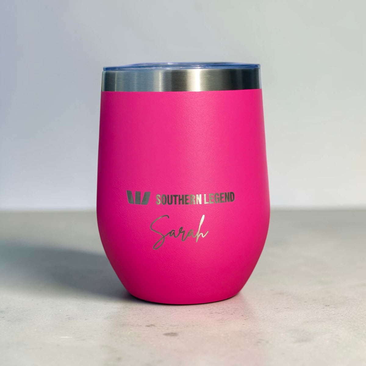 A bright pink insulated stemless tumbler with a brushed stainless steel rim and a clear lid, featuring the engraved text 'SOUTHEEN LEGEND' and the name 'Sarah' in silver on the front, set against a neutral grey background.