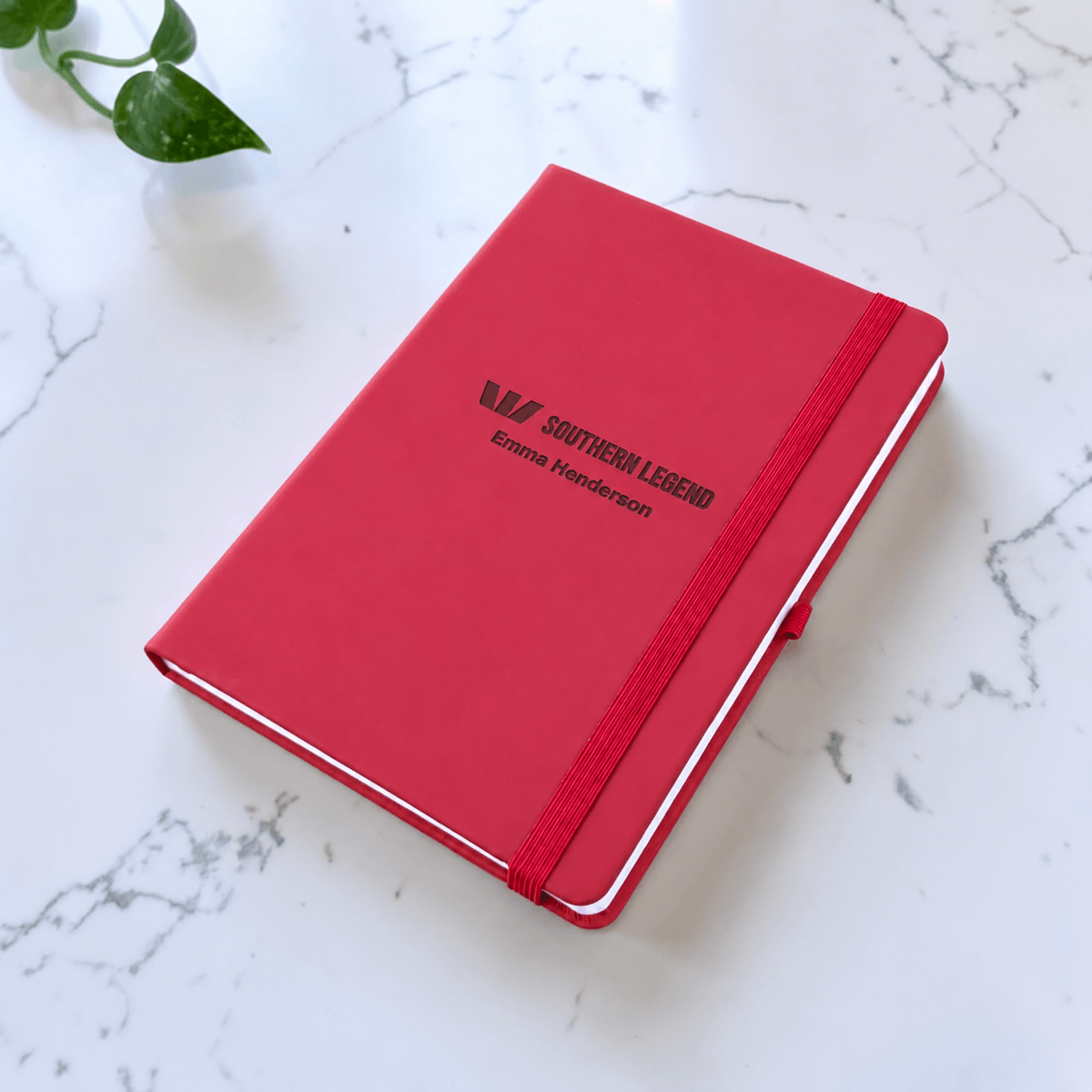A maroon hardcover notebook with an elastic closure band and engraved text reading 'SOUTHERN LEGEND Emma Henderson' on a white marble surface.