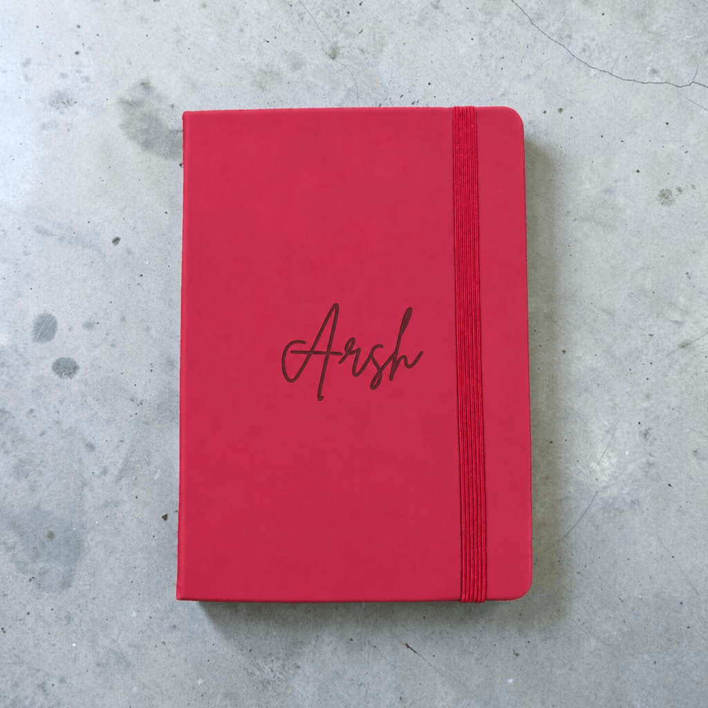 A maroon hardcover notebook with an elastic closure band and the name 'Arsh' engraved in cursive on the cover, placed on a gray concrete surface.