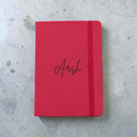 A maroon hardcover notebook with an elastic closure band and the name 'Arsh' engraved in cursive on the cover, placed on a gray concrete surface.