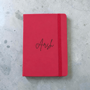 A maroon hardcover notebook with an elastic closure band and the name 'Arsh' engraved in cursive on the cover, placed on a gray concrete surface.