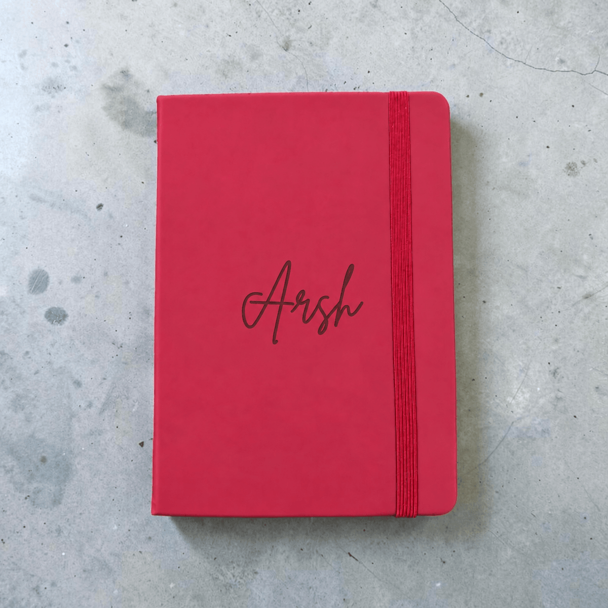 A maroon hardcover notebook with an elastic closure band and the name 'Arsh' engraved in cursive on the cover, placed on a gray concrete surface.