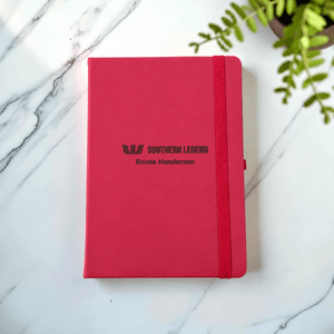 A maroon hardcover notebook with an elastic closure band, personalised with the text 'SOUTHERN LEGEND' and 'Emma Henderson', placed on a white marble surface with a green plant leaf in the corner.