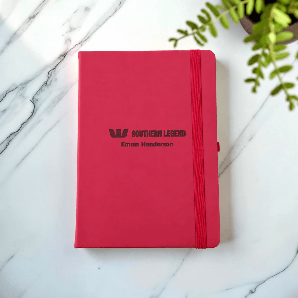 A maroon hardcover notebook with an elastic closure band, personalised with the text 'SOUTHERN LEGEND' and 'Emma Henderson', placed on a white marble surface with a green plant leaf in the corner.
