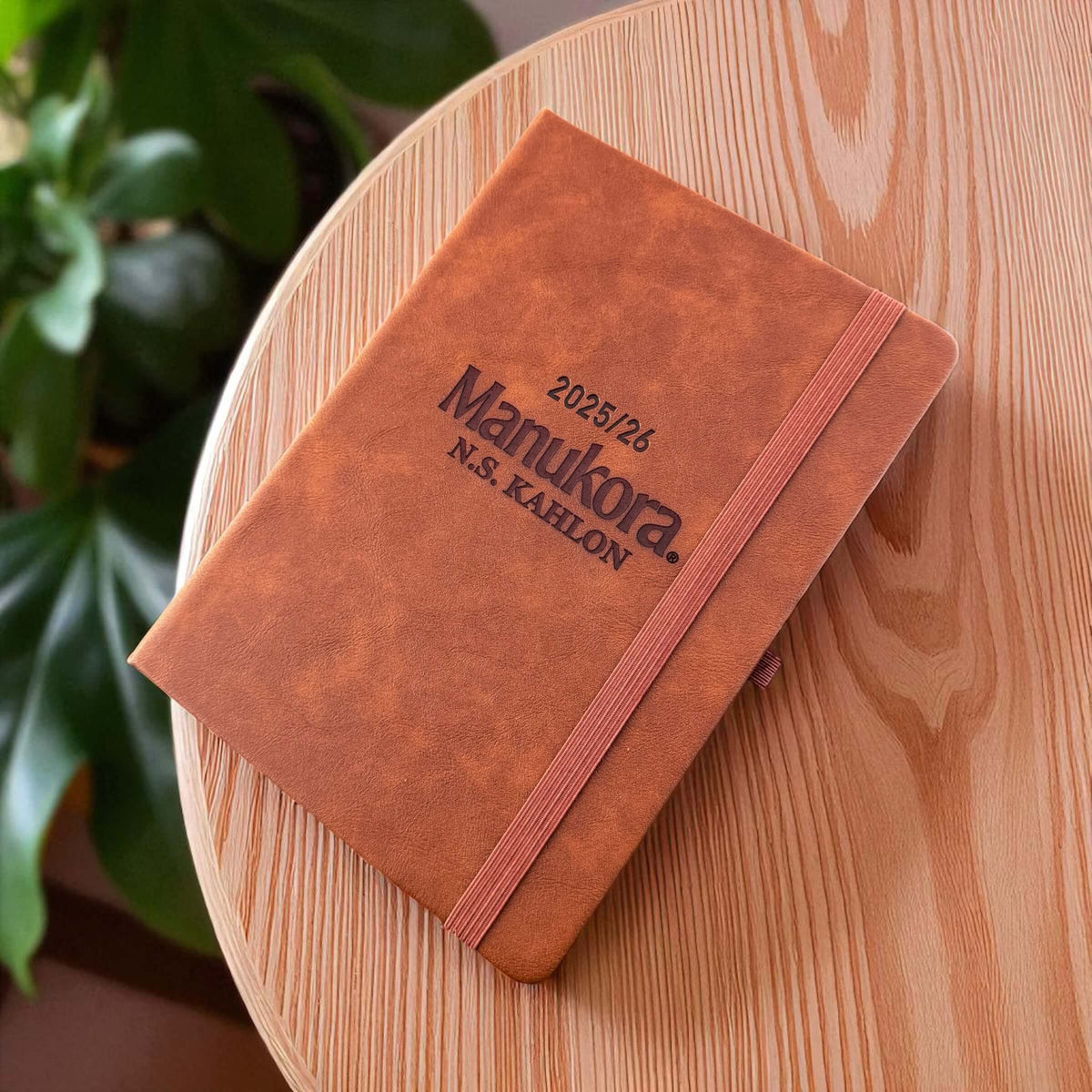 Angled overhead shot of the engraved business planner on a round timber table, showing elegant tan finish and elastic closure