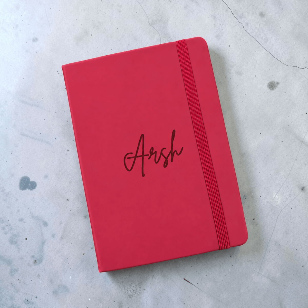 Angled view of an A5 maroon leather journal personalised with the name Arsh, showing the elastic closure, placed on a light gray concrete surface.