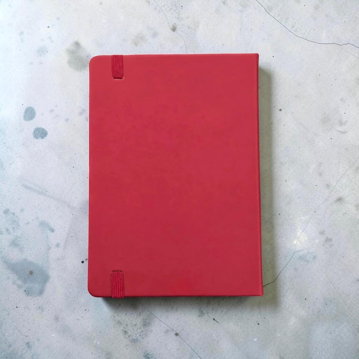 Back view of a maroon PU leather A5 notebook with a matching Maroon elastic closure tab, placed on a light gray concrete surface.