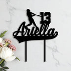 Black acrylic birthday cake topper shaped with a baseball player silhouette, custom name "Ariella" and age 13, styled on marble with white florals.