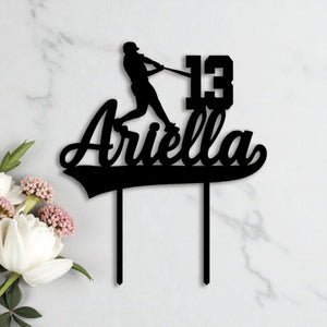Black acrylic birthday cake topper shaped with a baseball player silhouette, custom name "Ariella" and age 13, styled on marble with white florals.