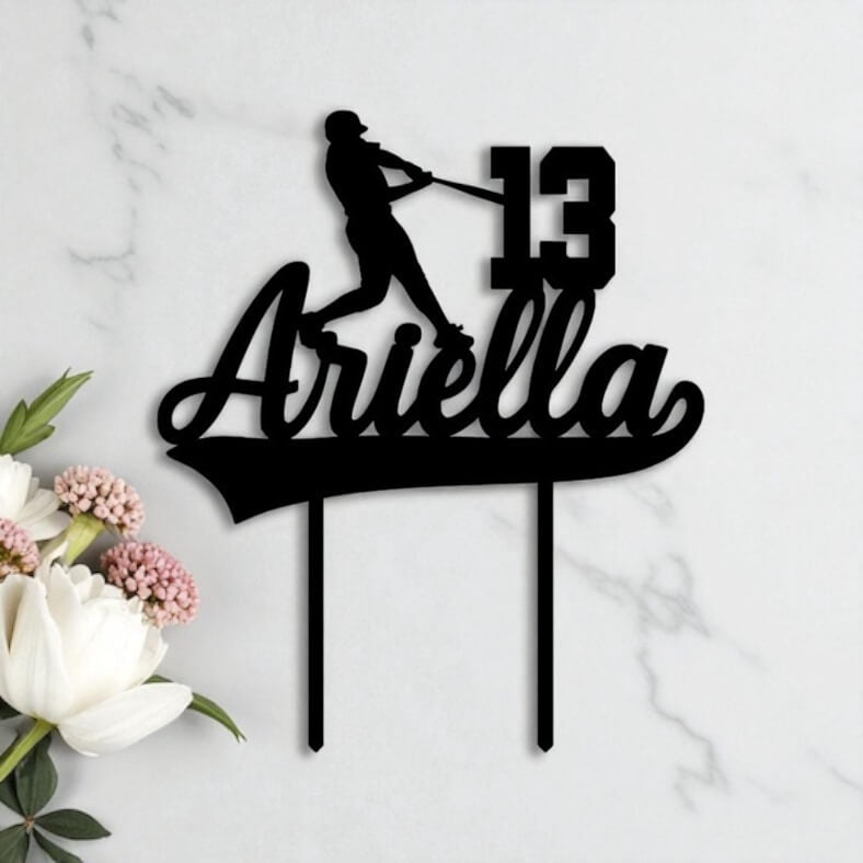 Black acrylic birthday cake topper shaped with a baseball player silhouette, custom name "Ariella" and age 13, styled on marble with white florals.