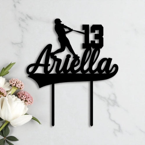 Black acrylic birthday cake topper shaped with a baseball player silhouette, custom name "Ariella" and age 13, styled on marble with white florals.