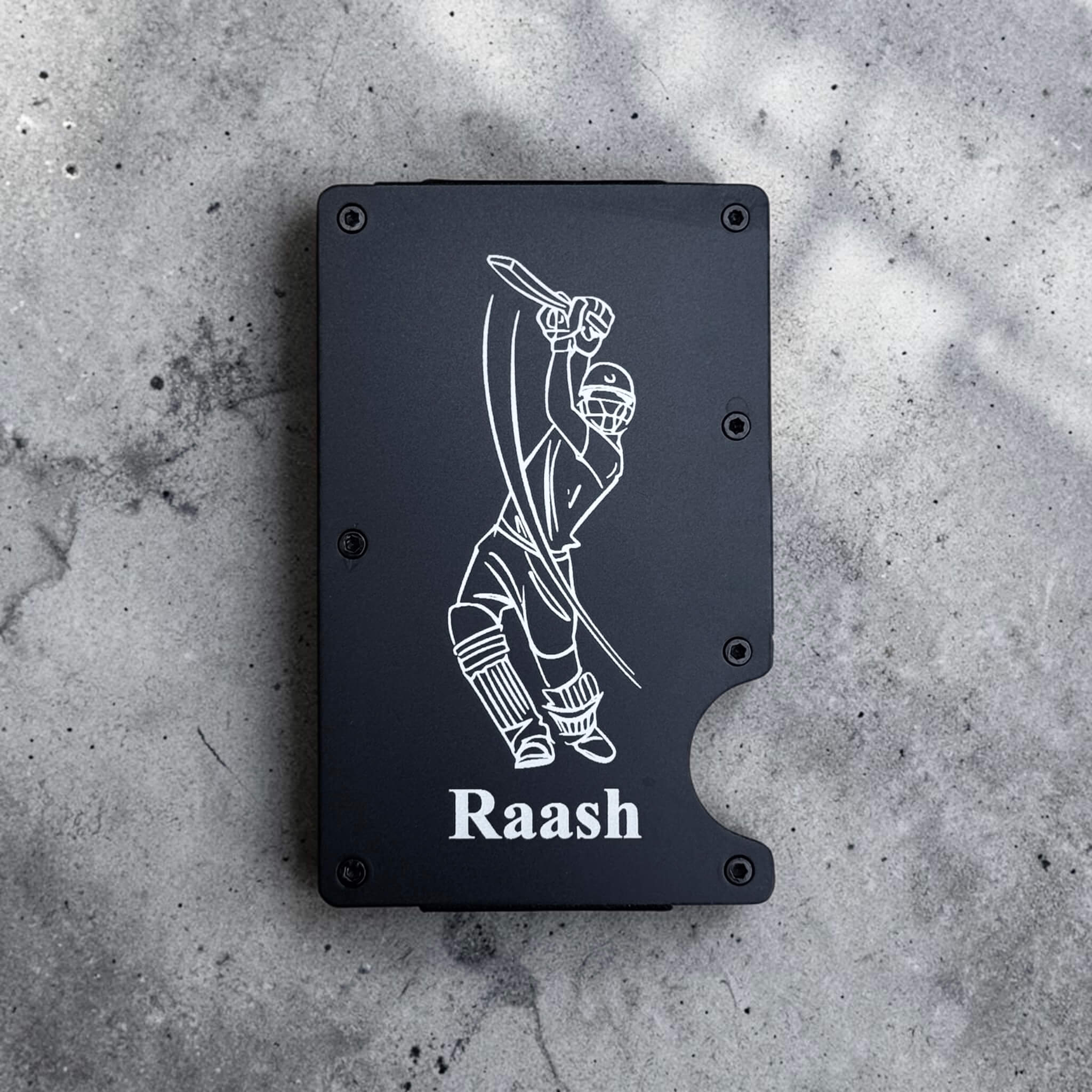 Black custom engraved metal card holder with personalised design and 'Raash' text on a gray concrete surface