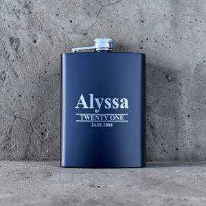 Black engraved birthday hip flask with name and date on a concrete surface