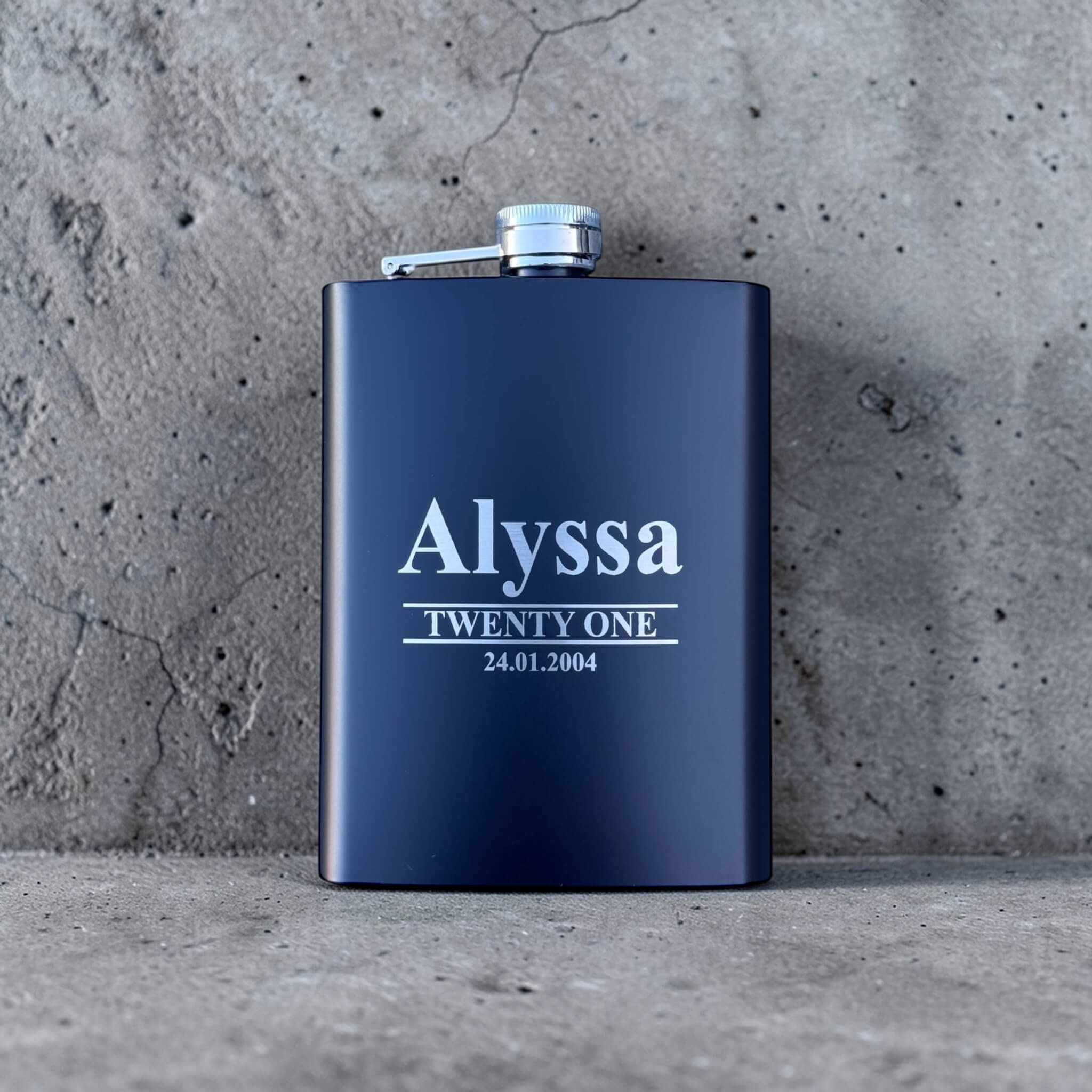 Black engraved birthday hip flask with name and date on a concrete surface