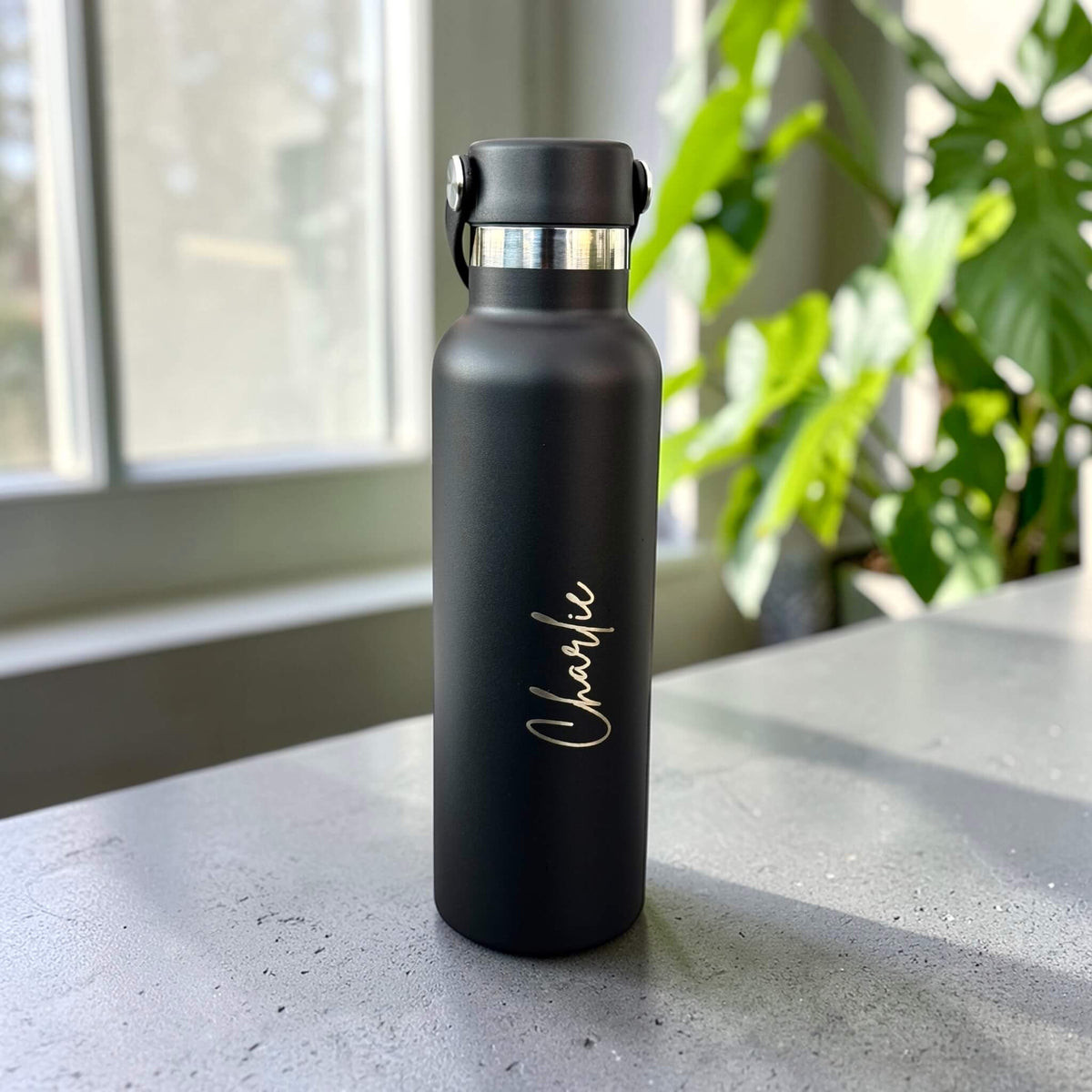 Black engraved water bottle with vertical script lettering and its carry handle folded down on a grey benchtop.