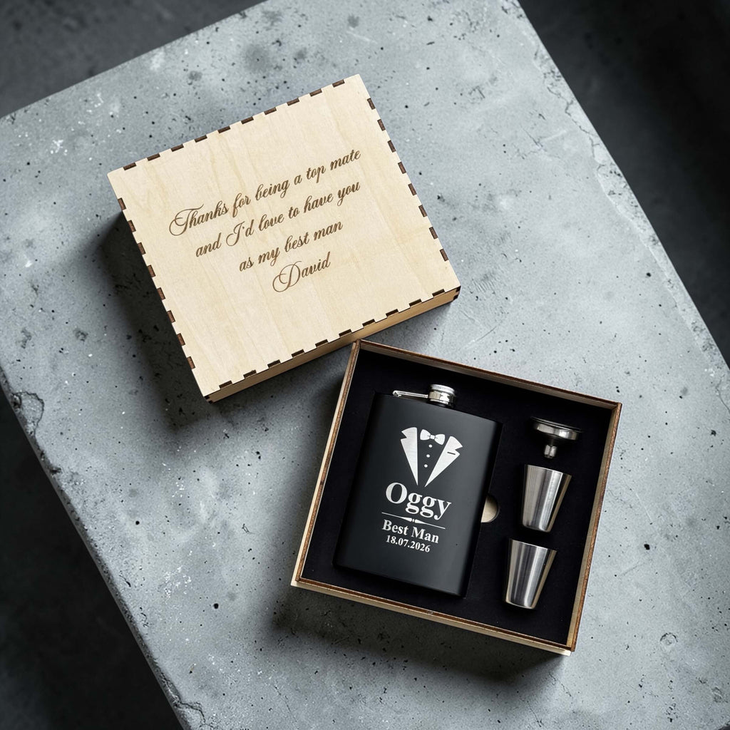 Black flask for wedding role & tuxedo design and 'Oggy Best Man' engraving, accompanied by two silver cups, in a wooden box on a concrete surface.