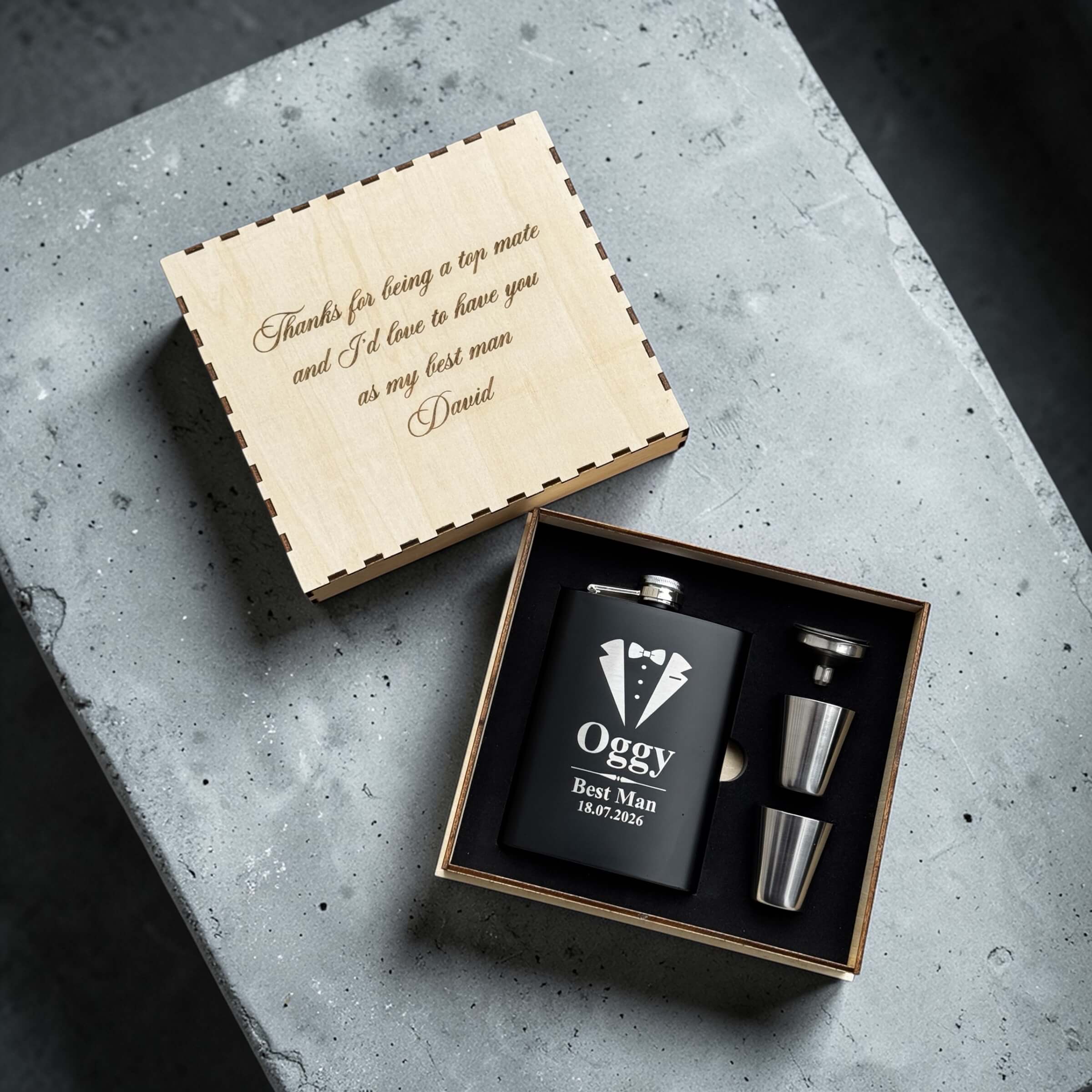 Black flask for wedding role & tuxedo design and 'Oggy Best Man' engraving, accompanied by two silver cups, in a wooden box on a concrete surface.