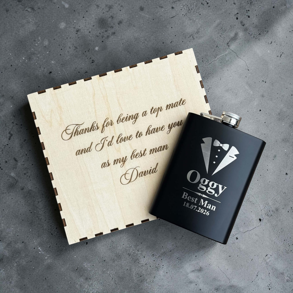 Black flask with 'Oggy Best Man' engraving and wooden gift box on a gray surface