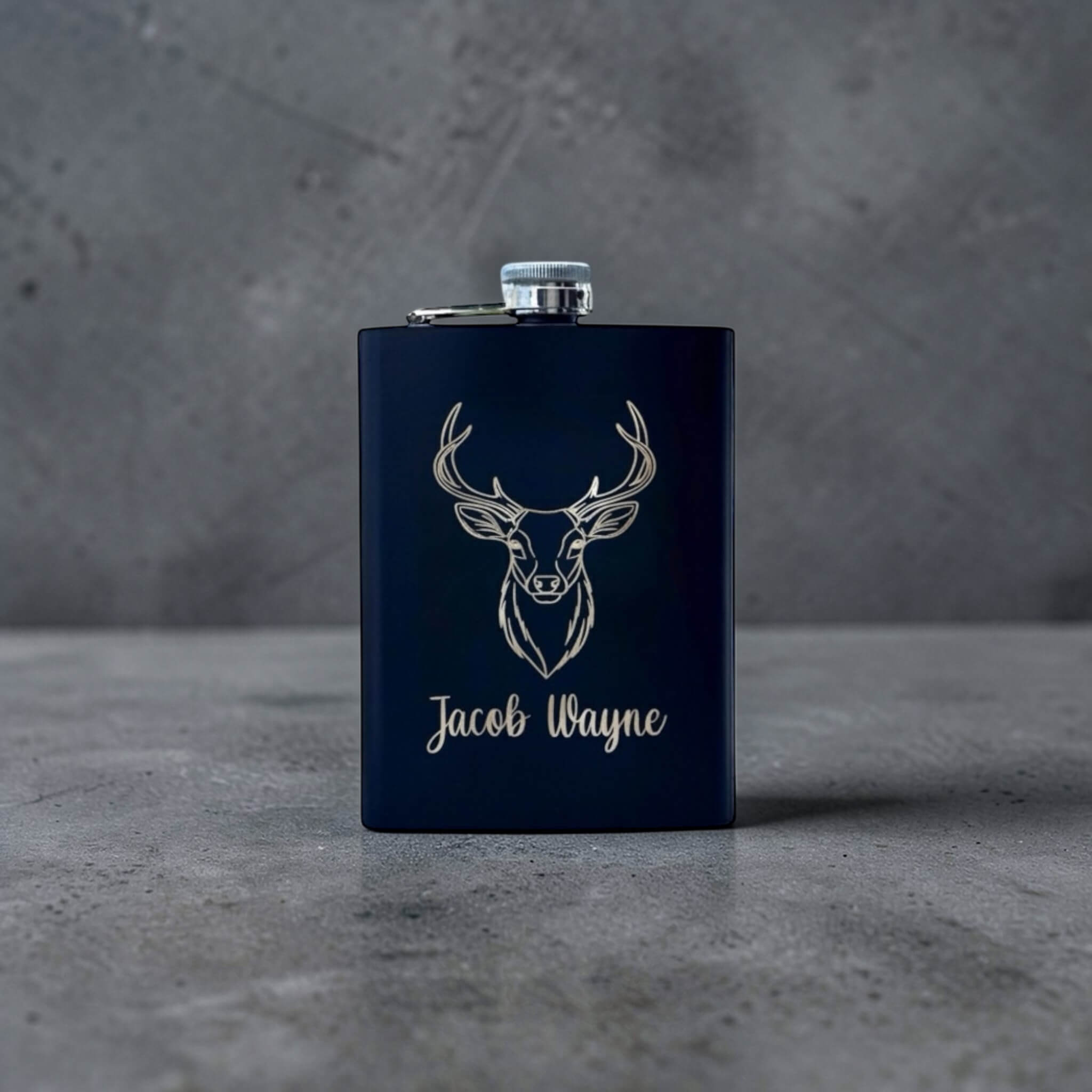 Black flask with deer head design and 'Jacob Wayne' text engraved on a gray surface.