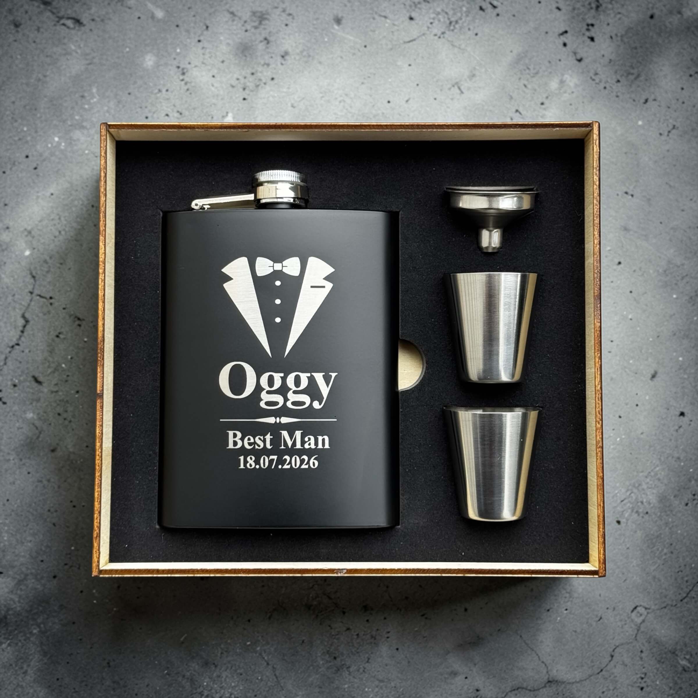 Black hip flask with tuxedo design and personalised text in a wooden box on a gray concrete background