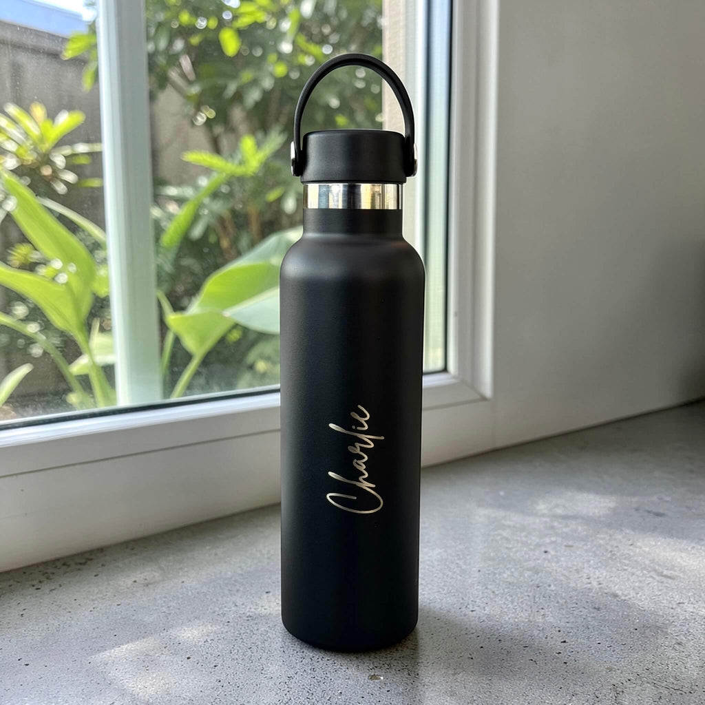 Black stainless steel water bottle engraved with Charlie in script, with its carry handle raised, beside a window.
