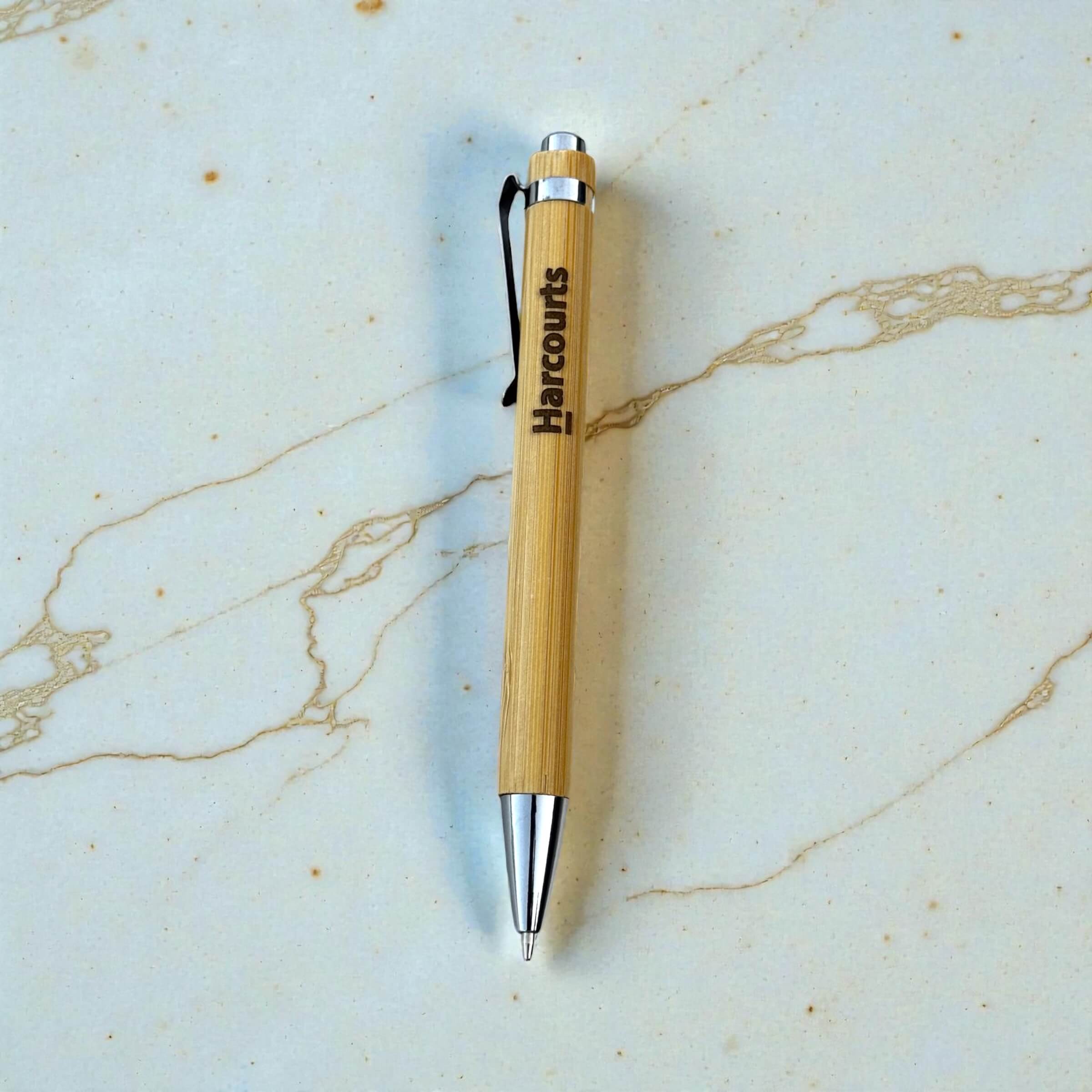 Branded bamboo ballpoint pen with laser engraved Harcourts logo, silver chrome tip and clip, on marble surface
