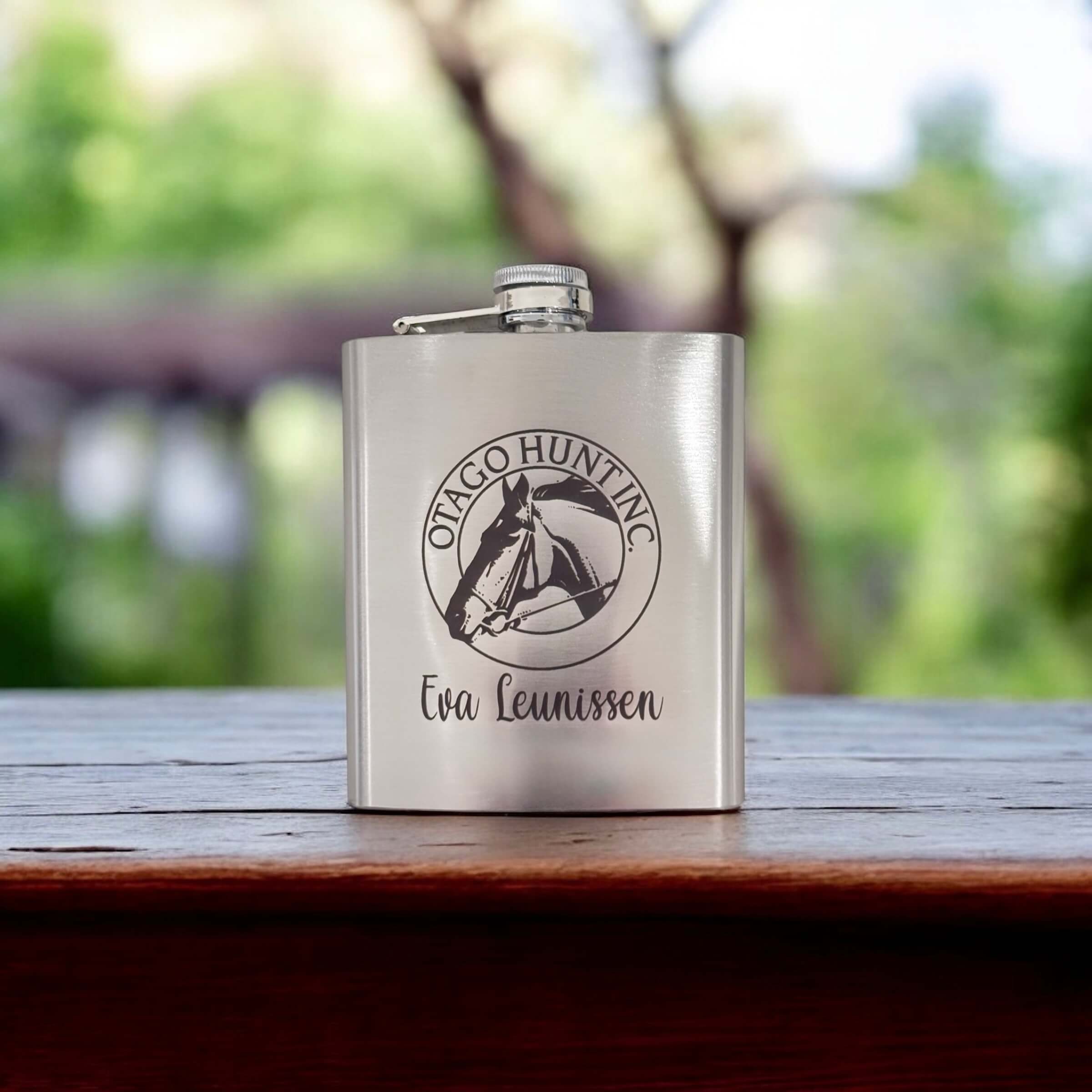 Branded stainless steel hip flask with engraved logo and personalised name, set against blurred greenery.