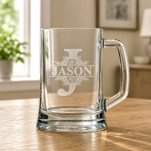 Clear glass beer mug with engraved Jason split monogram and floral detail on wooden table