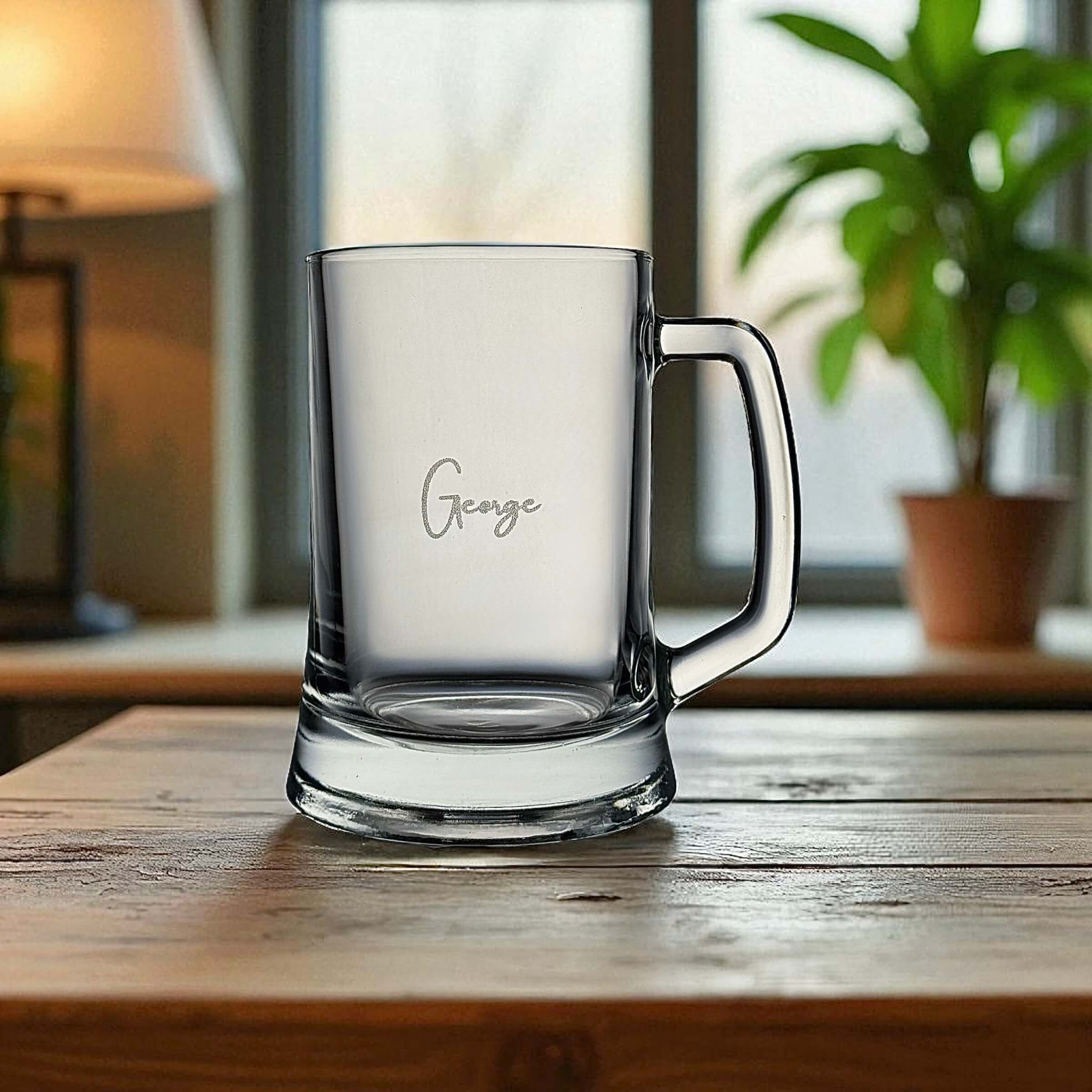 Clear glass mug with 'George' engraving on a wooden surface, blurred indoor background