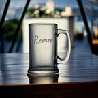 Clear glass mug with 'Romeo' engraving on a reflective surface with blurred background