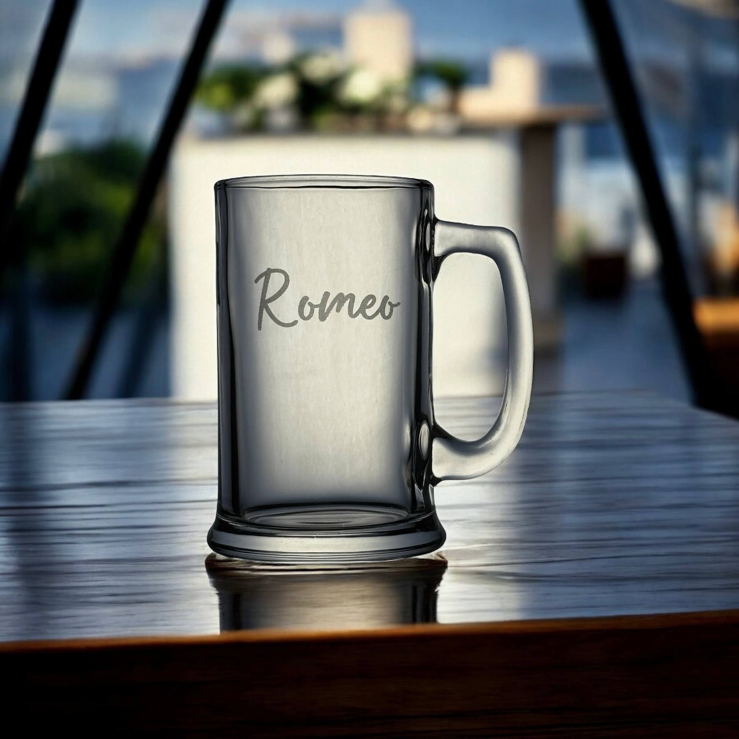 Clear glass mug with 'Romeo' engraving on a reflective surface with blurred background