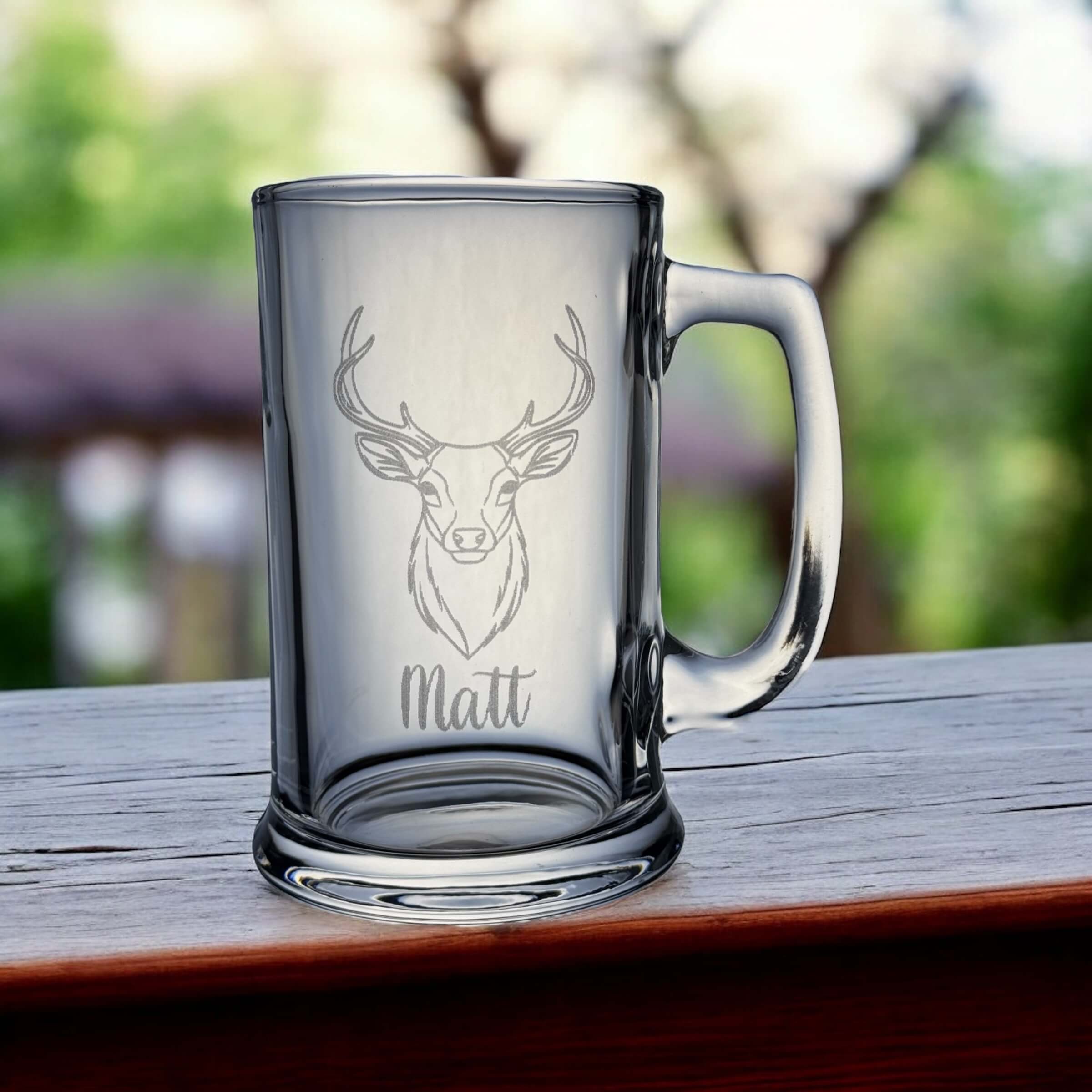 Clear glass mug with deer engraving and 'Matt' text on a wooden surface with blurred greenery in the background