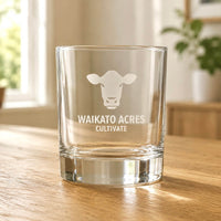 Clear whisky glass with engraved Waikato Acres cow logo on wooden table