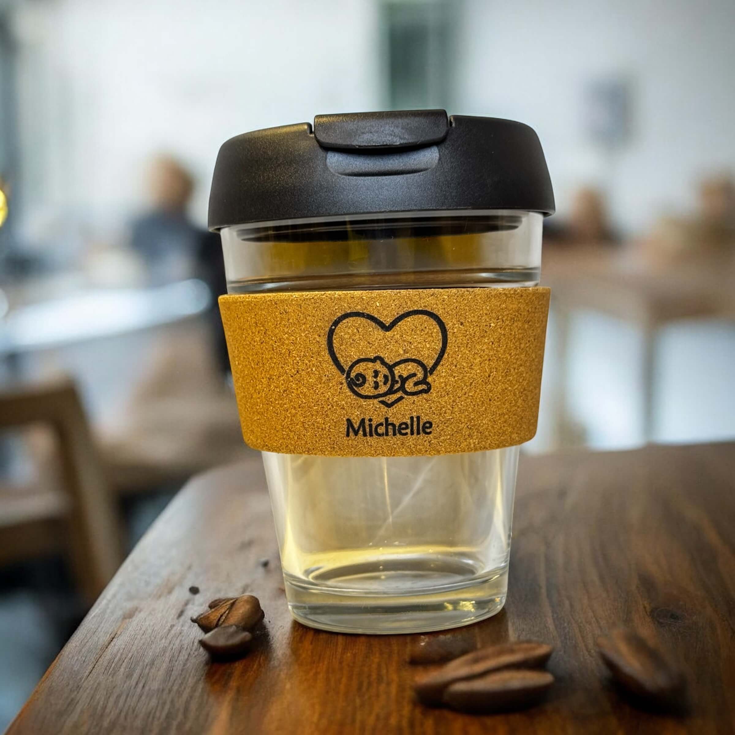 Close-up of a personalised glass coffee cup featuring the name Michelle on a cork sleeve, placed on a rustic wooden table with scattered coffee beans in a blurred coffee shop background