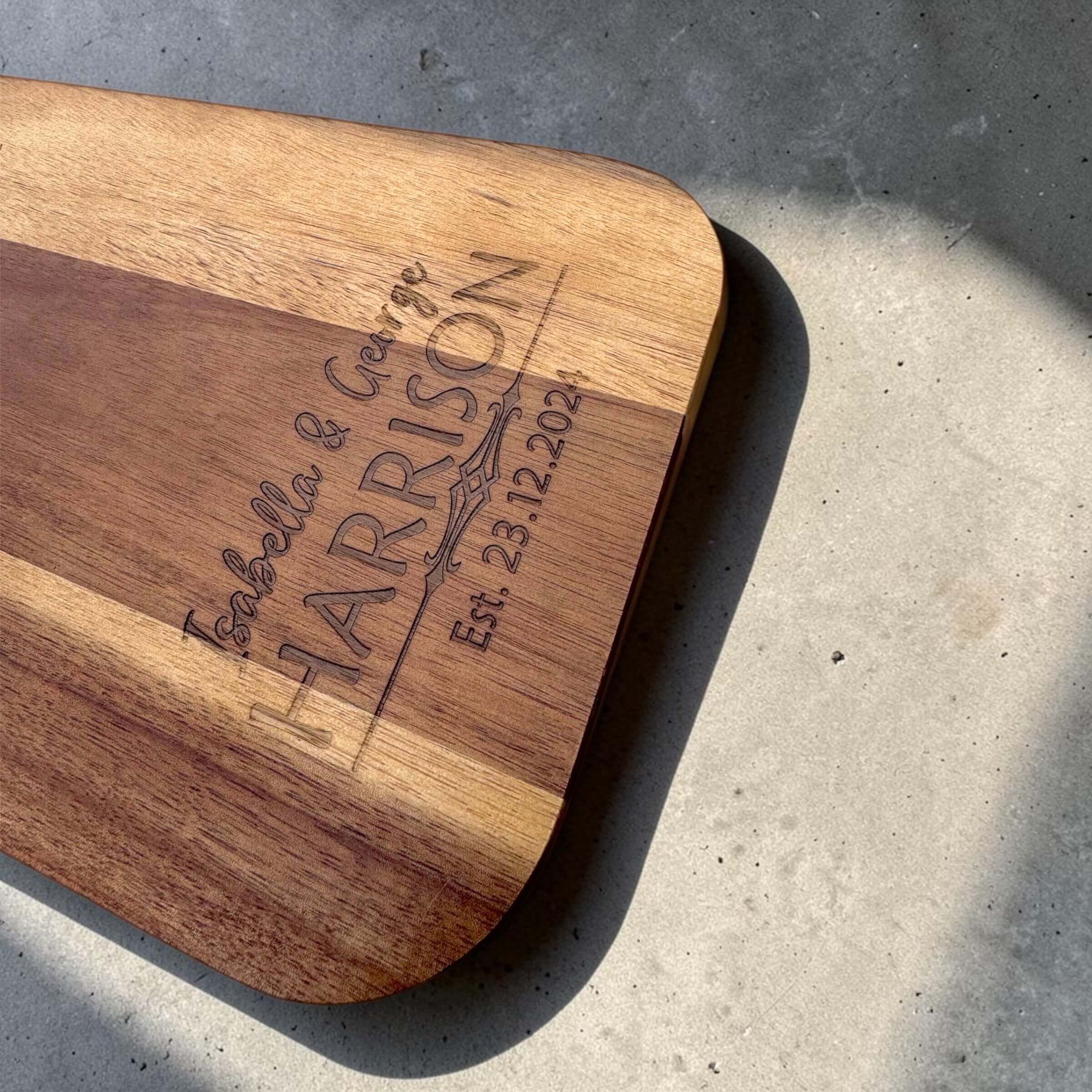 Close-up of fine laser engraving on an acacia wood serve board showing a couple's names, surname, and wedding date with natural wood grain contrast