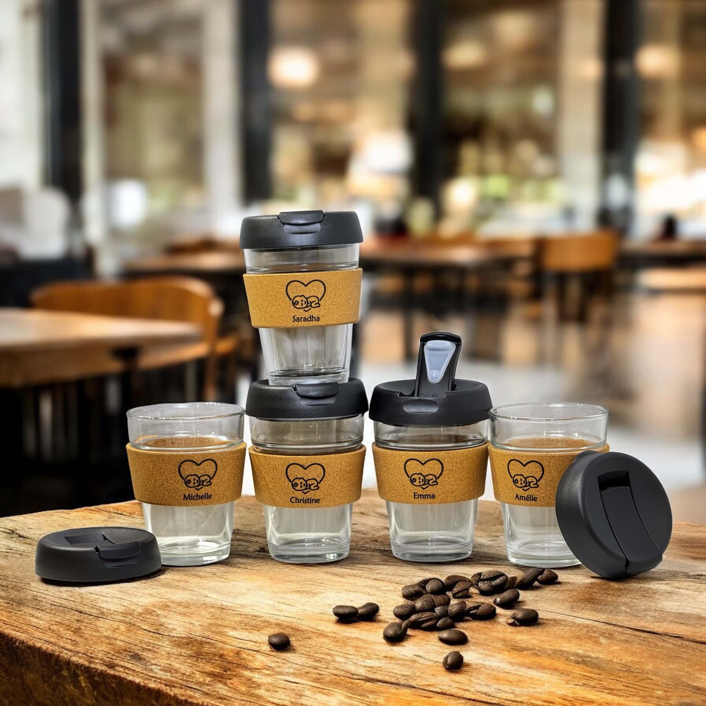 Collection of five reusable glass coffee cups with cork bands and custom names Saradha, Michelle, Christine, Emma and Amélie, arranged on a wooden table with coffee beans and lids in a cosy café backdrop