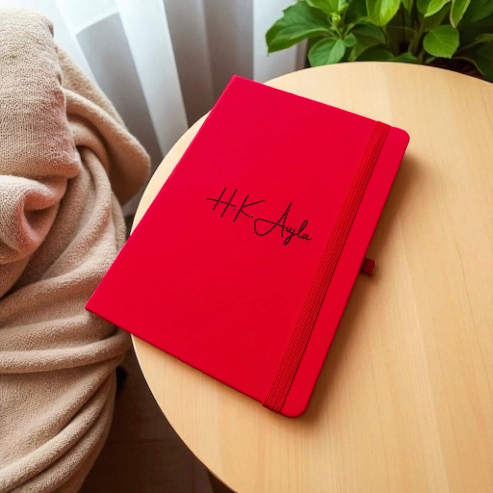 Custom engraved Red notebook with 'H.K. Aujla' branding on a wooden surface with a beige blanket and plant in the background.