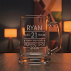 Custom glass beer stein engraved “Ryan 21st Birthday Brewing Since 2004” on dark wooden table in cosy room