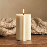 Custom scented white ribbed candle with a lit wick on a wooden surface with a beige knitted blanket in the background.