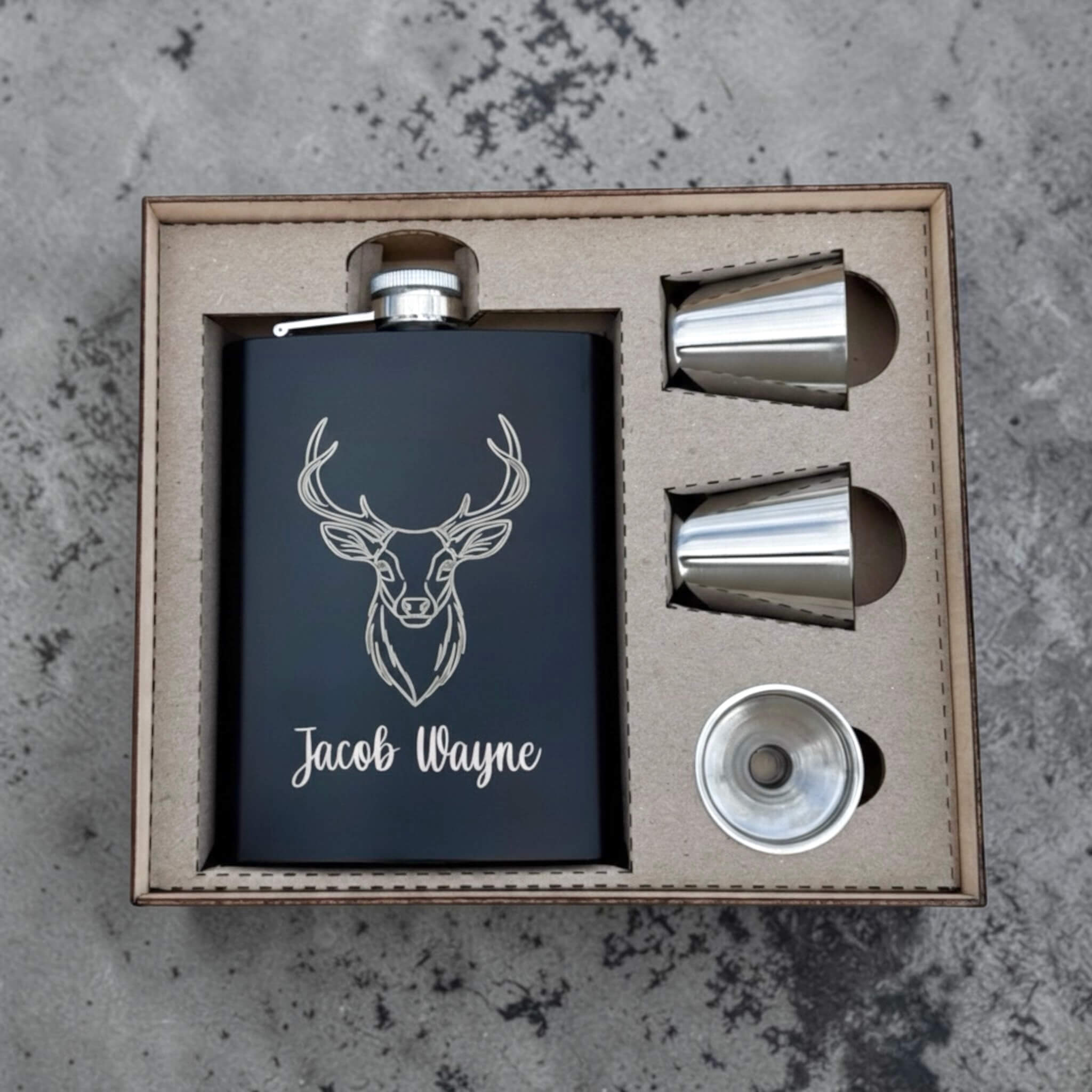 Flask set with deer head design and 'Jacob Wayne' text engraved in a wooden box on a textured surface.
