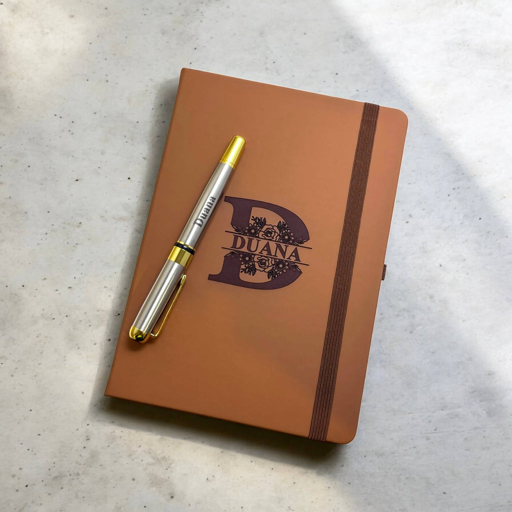 Flat-lay of personalised notebook featuring split floral initial and engraved pen placed diagonally on the cover