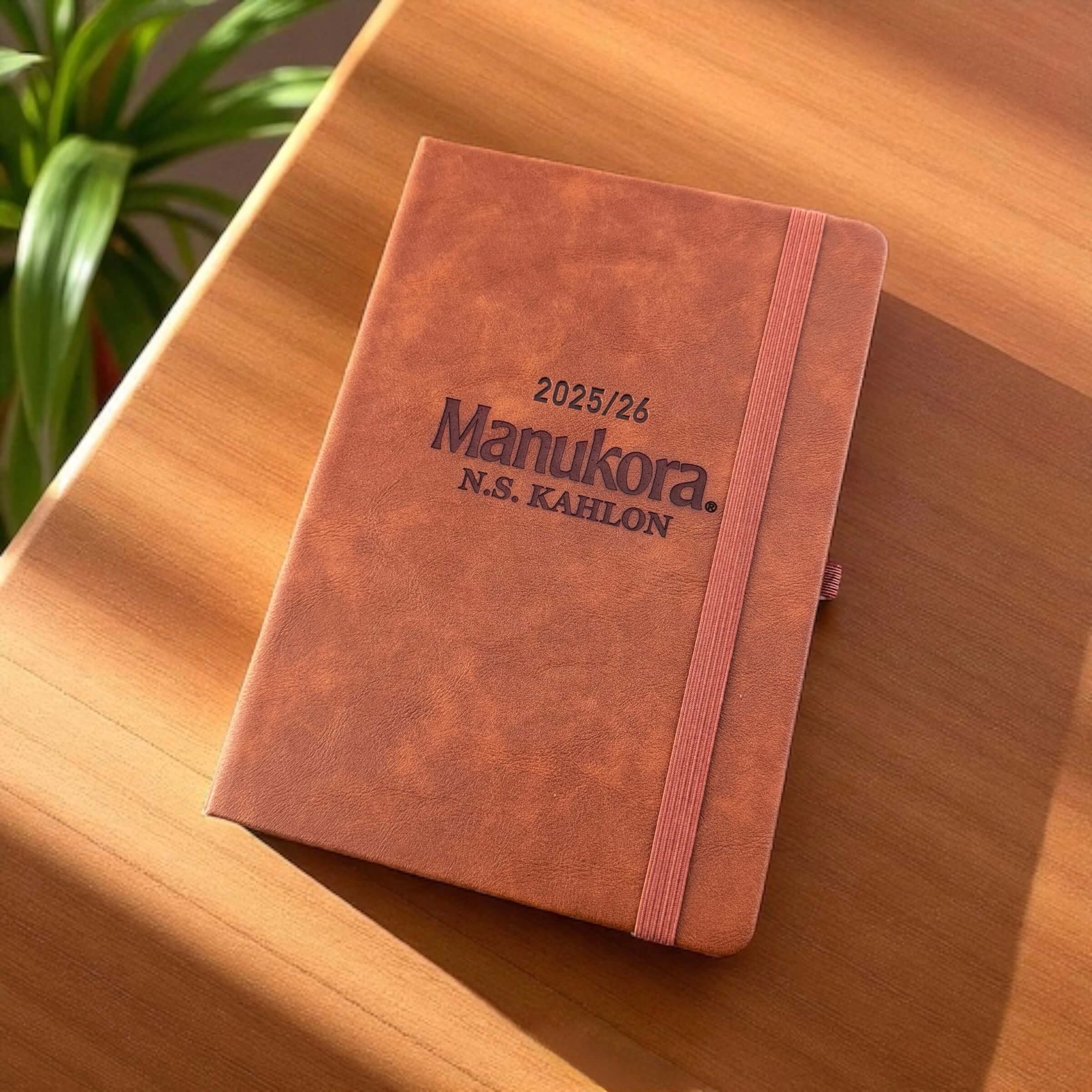 Flat-lay of the engraved leather planner on a sunlit desk, highlighting the bold Manukora branding and professional design