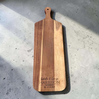 Flat lay of a long-handled acacia wood serve board laser engraved with a couple's first names and wedding date, photographed on concrete in natural light