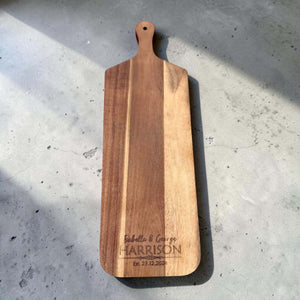 Flat lay of a long-handled acacia wood serve board laser engraved with a couple's first names and wedding date, photographed on concrete in natural light