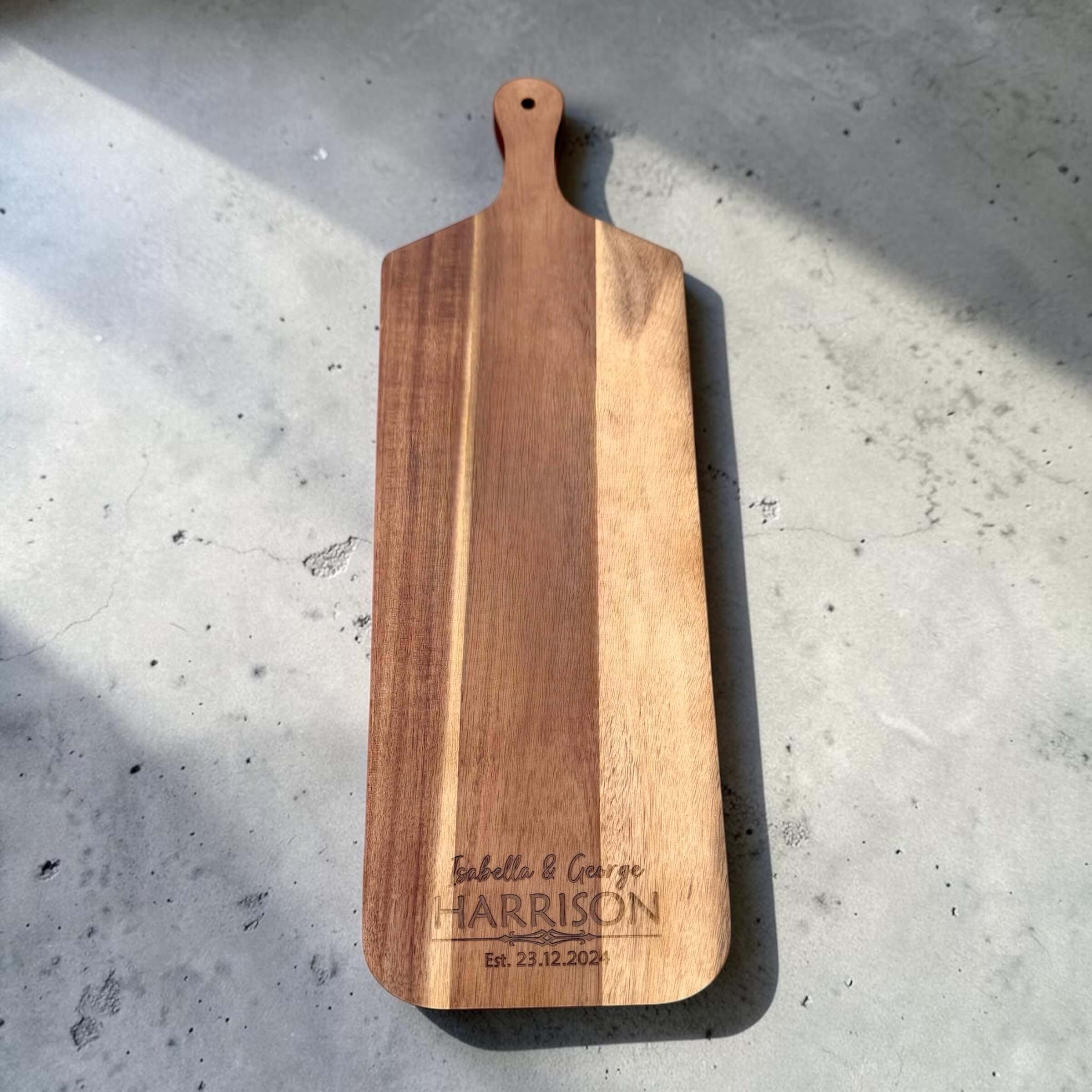 Flat lay of a long-handled acacia wood serve board laser engraved with a couple's first names and wedding date, photographed on concrete in natural light